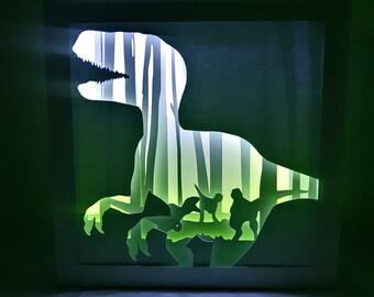 Pode incluir: Uma peça de arte em papel em camadas com uma silhueta de dinossauro com uma cena de floresta no interior. A obra de arte é iluminada com luz verde e branca, criando um efeito dramático. A moldura é branca.