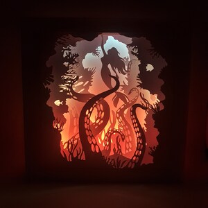 May include: A paper cut-out art piece depicting a silhouette of a woman with long hair, surrounded by the tentacles of an octopus. The artwork is illuminated from behind, creating a glowing effect.