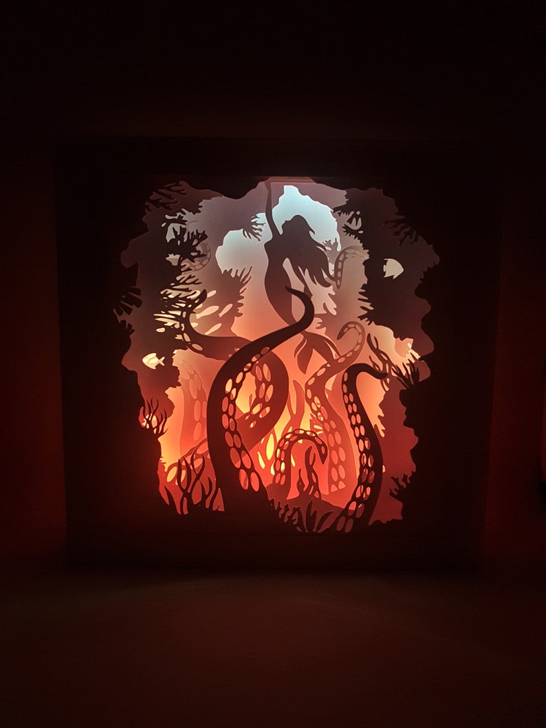 May include: A paper cut-out art piece depicting a silhouette of a woman with long hair, surrounded by the tentacles of an octopus. The artwork is illuminated from behind, creating a glowing effect.