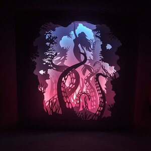 May include: A paper cut-out art piece depicting a mermaid being pulled into the depths by an octopus. The artwork is illuminated from behind with pink and blue lights, creating a dramatic effect.