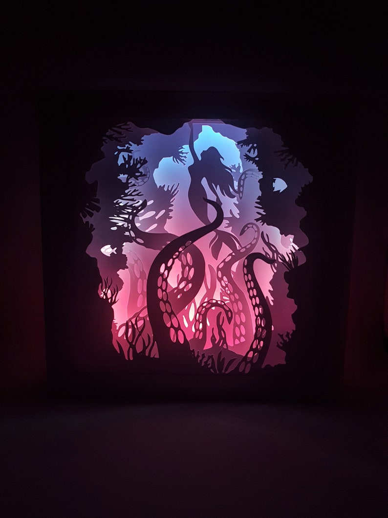 May include: A paper cut-out art piece depicting a mermaid being pulled into the depths by an octopus. The artwork is illuminated from behind with pink and blue lights, creating a dramatic effect.