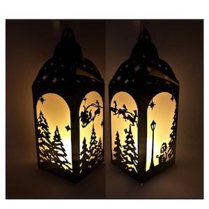 May include: A black metal lantern with a white paper insert depicting a Christmas scene. The scene shows a silhouette of Santa Claus flying in a sleigh pulled by reindeer, a snowman, and a street lamp. The lantern is lit from within, casting a warm glow.