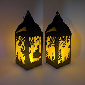 Könnte beinhalten: Zwei schwarze Papierlaternen mit einem ausgeschnittenen Design einer Person auf einer Schaukel, die unter einem Baum liest. Die Laternen sind mit warmem gelbem Licht beleuchtet und schaffen eine gemütliche Atmosphäre.