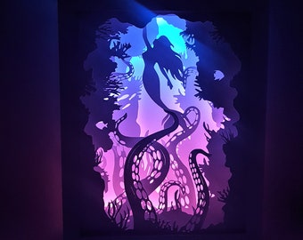 May include: A layered paper art piece depicting a mermaid swimming in an underwater scene with tentacles and aquatic plants. The artwork is illuminated with blue, purple, and pink lights, creating a dramatic effect.