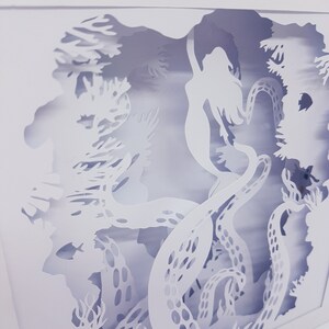 May include: White paper cut-out art depicting a mermaid swimming through an underwater scene with an octopus and coral. The artwork is framed in a white square.