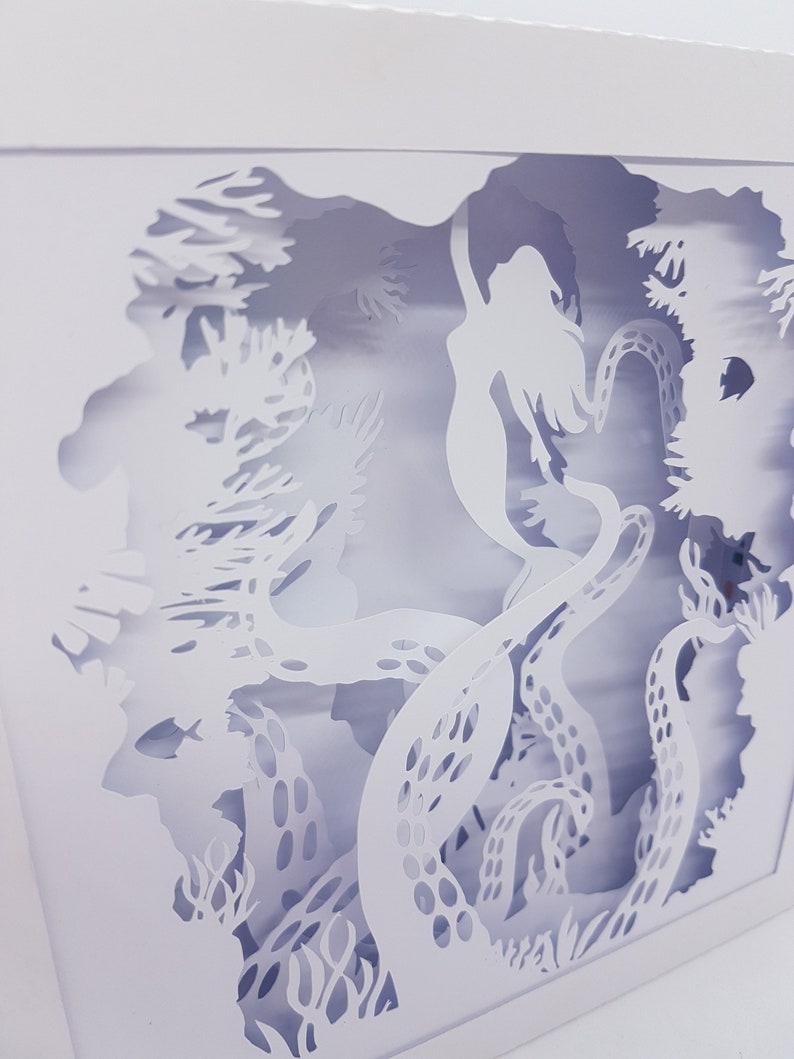 May include: White paper cut-out art depicting a mermaid swimming through an underwater scene with an octopus and coral. The artwork is framed in a white square.