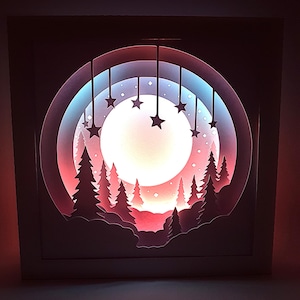 Könnte beinhalten: Ein geschichtetes Papierkunstwerk, das eine nächtliche Waldszene darstellt. Das Kunstwerk zeigt einen großen, leuchtenden Mond, silhouettenartige Bäume und hängende Sternformen. Die Farbpalette umfasst Rot-, Blau- und Weißtöne und erzeugt einen warmen, beleuchteten Effekt.