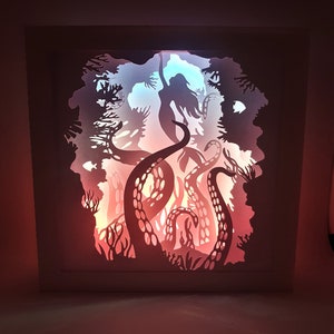 May include: A white papercraft lamp with a silhouette of a mermaid being pulled down by an octopus's tentacles. The lamp is lit from behind, casting a warm glow through the cutouts.