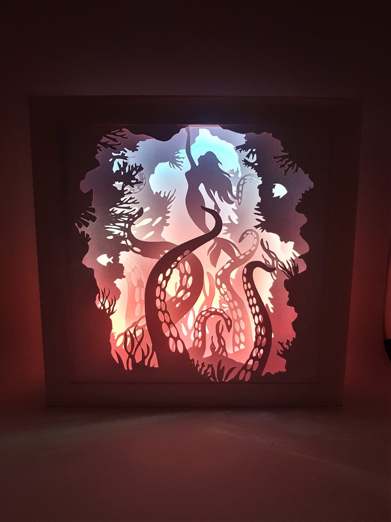 May include: A white papercraft lamp with a silhouette of a mermaid being pulled down by an octopus's tentacles. The lamp is lit from behind, casting a warm glow through the cutouts.