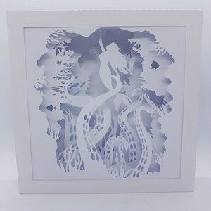 May include: White paper cut-out art depicting a mermaid surrounded by an octopus's tentacles. The artwork is framed in a white frame.