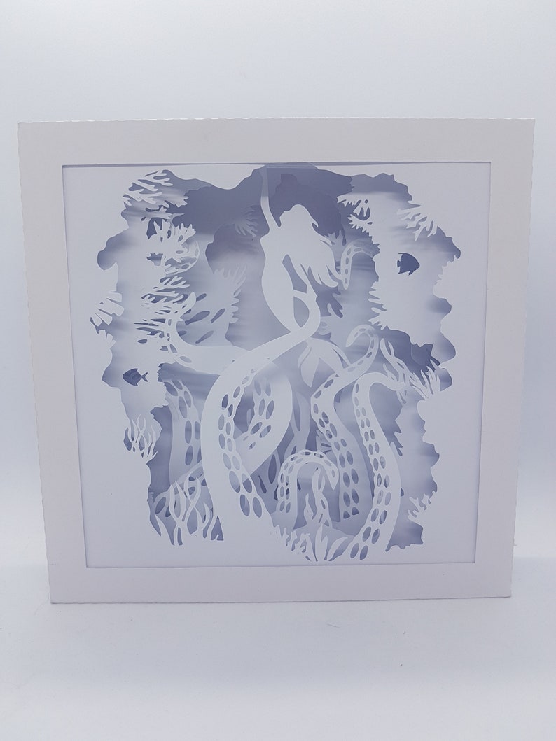 May include: White paper cut-out art depicting a mermaid surrounded by an octopus's tentacles. The artwork is framed in a white frame.
