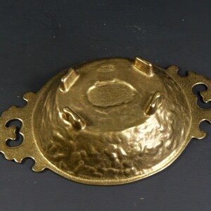May include: A gold-toned metal serving dish with a hammered finish and ornate handles. The dish has three small feet on the bottom.