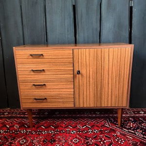 Könnte beinhalten: Ein moderner Holzschrank aus der Mitte des Jahrhunderts mit vier Schubladen und einer einzigen Tür. Der Schrank hat eine hellbraune Holzoptik mit einer Holzmaserung. Der Schrank steht auf einem roten Teppich mit Muster.