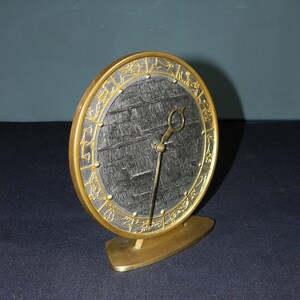 Puede incluir: Un reloj dorado con una esfera negra y detalles dorados. La esfera del reloj presenta un diseño de símbolos del zodíaco alrededor del borde. El reloj está sobre un soporte dorado.