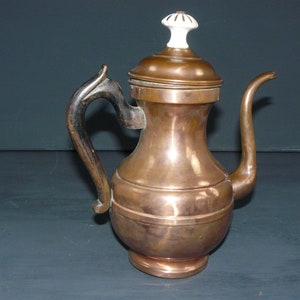 Op de afbeelding: Een koperen theepot met een zwarte houten handgreep en een witte porseleinen knop op de deksel.