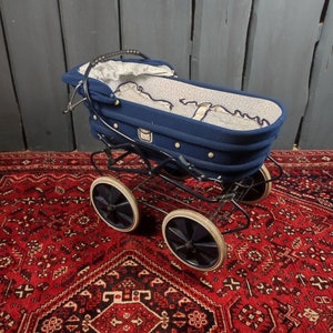 May include: A blue baby stroller with a white and blue patterned lining. The stroller has four black wheels with white tires and a black metal frame. The stroller is sitting on a red rug with a patterned design.