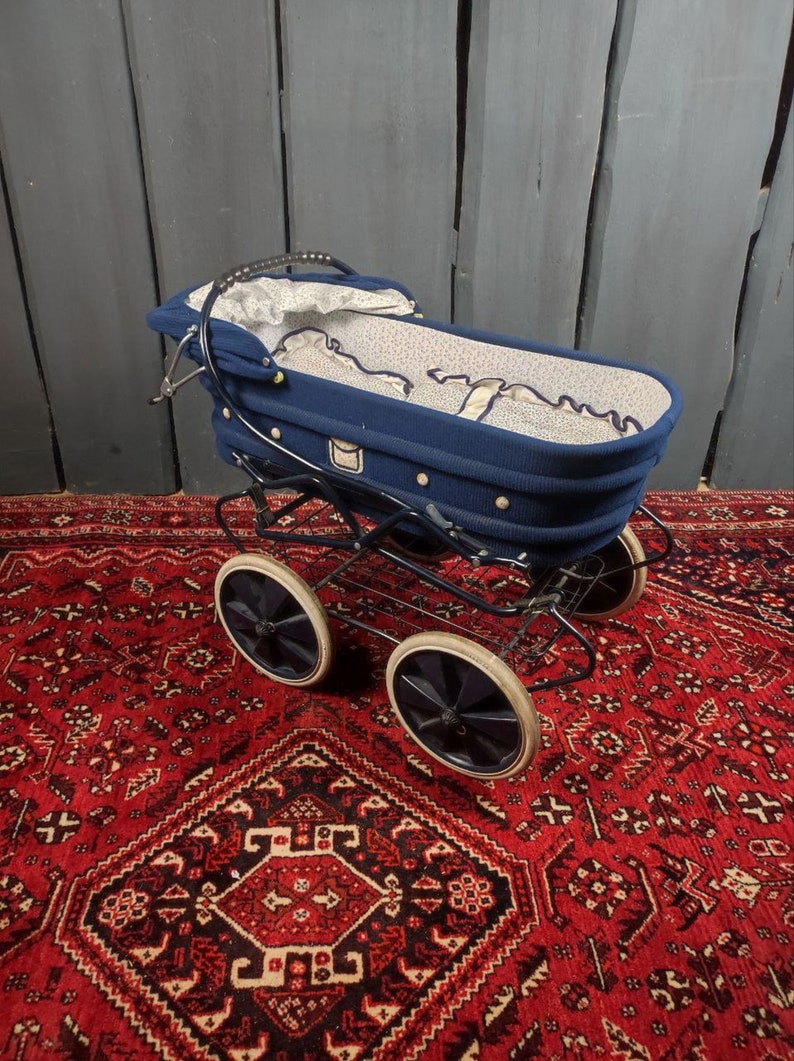 May include: A blue baby stroller with a white and blue patterned lining. The stroller has four black wheels with white tires and a black metal frame. The stroller is sitting on a red rug with a patterned design.