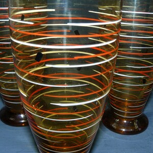 K&ouml;nnte beinhalten: Drei bernsteinfarbene Gl&auml;ser mit wei&szlig;en und orangefarbenen Spiralmustern. Die Gl&auml;ser haben einen klaren Boden.
