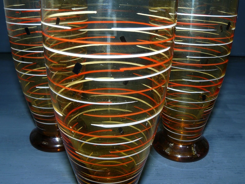 K&ouml;nnte beinhalten: Drei bernsteinfarbene Gl&auml;ser mit wei&szlig;en und orangefarbenen Spiralmustern. Die Gl&auml;ser haben einen klaren Boden.