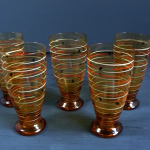 K&ouml;nnte beinhalten: Satz von f&uuml;nf Trinkgl&auml;sern aus Bernstein-Glas mit einem wei&szlig;en und orangefarbenen Wirbelmuster. Die Gl&auml;ser haben einen abgerundeten Boden und eine hohe, zylindrische Form.