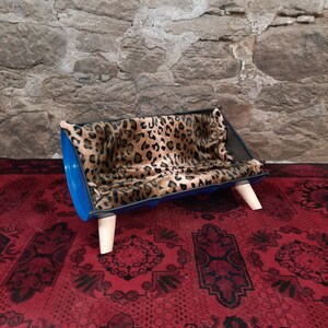 Op de afbeelding: Een blauw en zwart hondenbed met een kussen met luipaardprint. Het bed heeft houten poten en staat op een rood tapijt.
