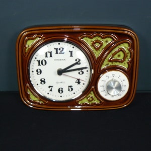 Może przedstawiać: Brązowy ceramiczny zegar ścienny z białą tarczą i mniejszą tarczą. Tarcza zegara ma czarne cyfry i wskazówki, z nazwą marki "DUGENA" wydrukowaną nad środkiem. Zegar ma zielone i brązowe dekoracyjne akcenty.