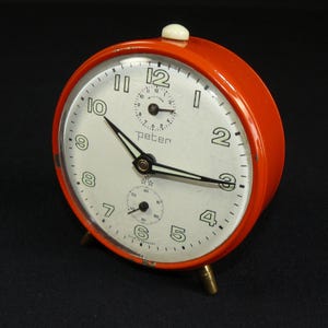 Könnte beinhalten: Ein roter Wecker mit weißem Zifferblatt und schwarzen Zeigern. Die Uhr hat ein kleines Sekundenzeiger-Zifferblatt und das Wort "Peter" ist auf dem Zifferblatt aufgedruckt.