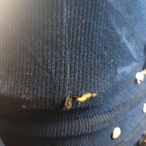 May include: A close-up of a dark blue corduroy fabric with a small tear and a yellow stain. The fabric has a ribbed texture.