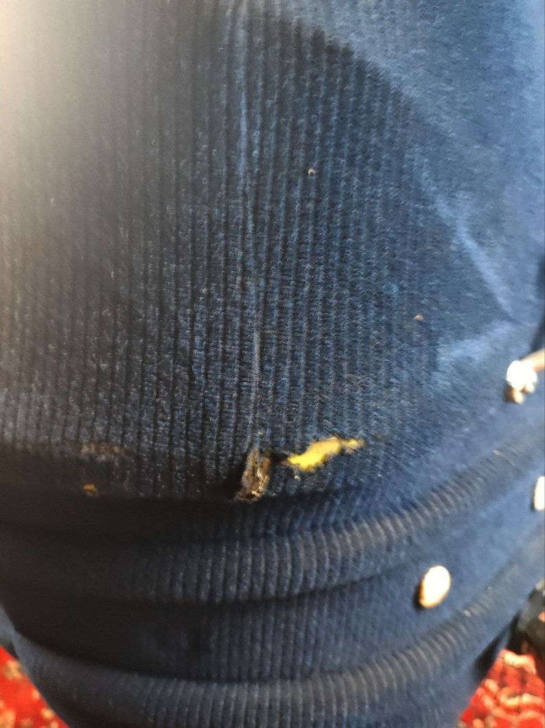 May include: A close-up of a dark blue corduroy fabric with a small tear and a yellow stain. The fabric has a ribbed texture.