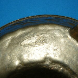 May include: A silver-colored metal bowl with a hammered texture and a circular logo on the bottom. The bowl has three feet and is designed for serving.