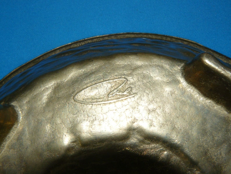 May include: A silver-colored metal bowl with a hammered texture and a circular logo on the bottom. The bowl has three feet and is designed for serving.