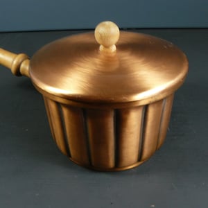 May include: A copper saucepan with a wooden handle and a lid. The saucepan has a fluted design and a wooden knob on the lid.