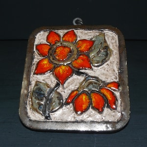 Könnte beinhalten: Ein Keramik-Wandbehang mit einem erhabenen Design aus zwei orangefarbenen und roten Blumen mit grünen Blättern auf weißem Hintergrund. Der Behang ist in einem braunen Rahmen gefasst.