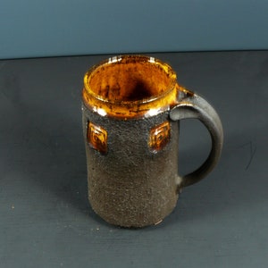 Könnte beinhalten: Eine braune Keramiktasse mit dunkelbrauner Glasur und gelbem Rand. Die Tasse hat einen Henkel und ein geometrisches Muster auf der Vorderseite.