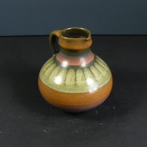 Könnte beinhalten: Ein kleiner, brauner Keramik Krug mit einem grün-orange-violetten Streifenmuster. Der Krug hat einen runden Körper und einen kleinen Henkel.