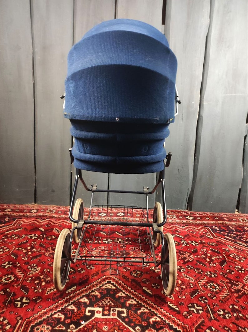 May include: A vintage blue stroller with a black metal frame and a basket underneath. The stroller has three wheels and is on a red patterned rug.