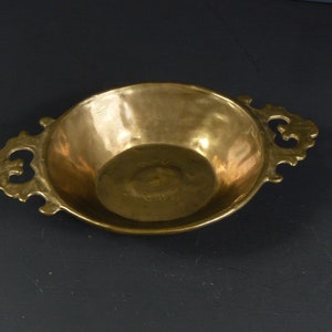 May include: A small, round, brass serving dish with two ornate handles. The dish has a smooth, polished surface and a slightly raised rim.
