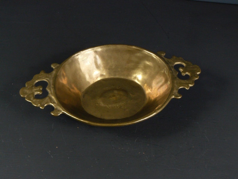May include: A small, round, brass serving dish with two ornate handles. The dish has a smooth, polished surface and a slightly raised rim.