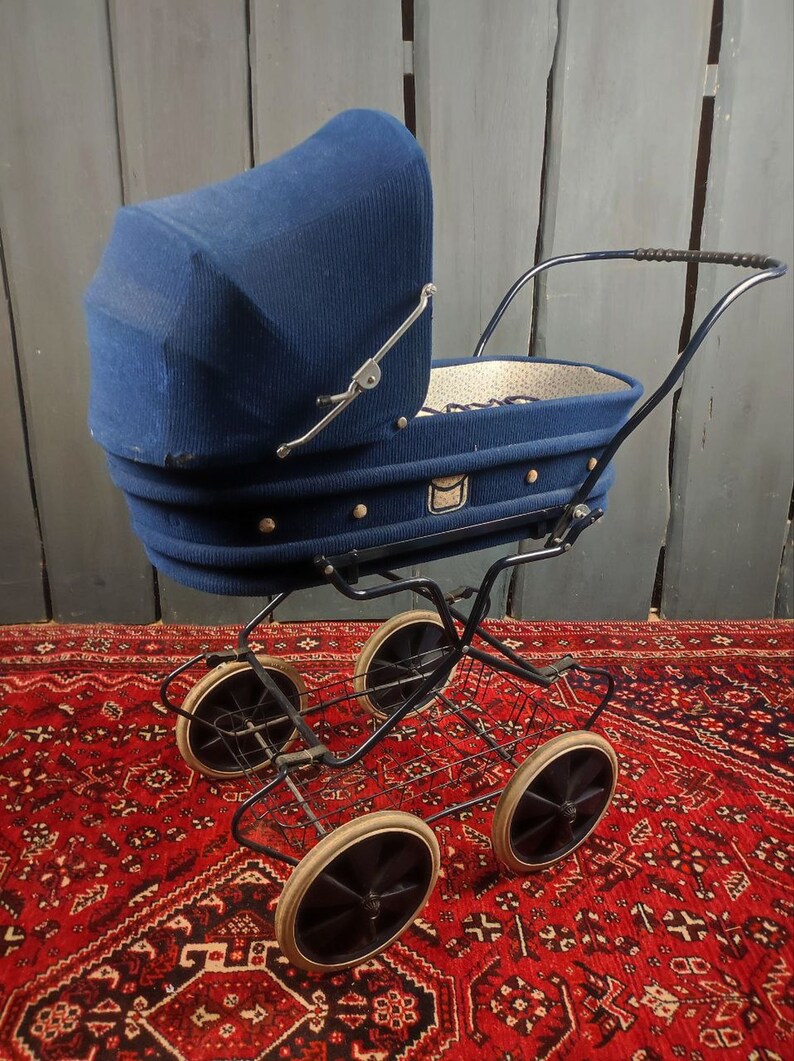 May include: A vintage blue baby stroller with a fabric canopy and a wire basket underneath. The stroller has four wheels and is sitting on a red patterned rug.