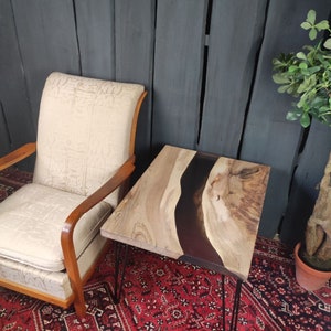 Op de afbeelding: Een vierkante houten tafel met een zwarte epoxyharsrivier die door het midden stroomt. De tafel heeft haarspeldpoten en staat op een rood tapijt.