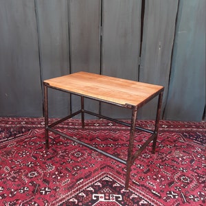 Könnte beinhalten: Ein kleiner Holztisch mit einem schwarzen Metallrahmen. Der Tisch hat eine rechteckige Tischplatte und ein Regal darunter. Der Tisch steht auf einem roten Teppich mit geometrischem Muster.