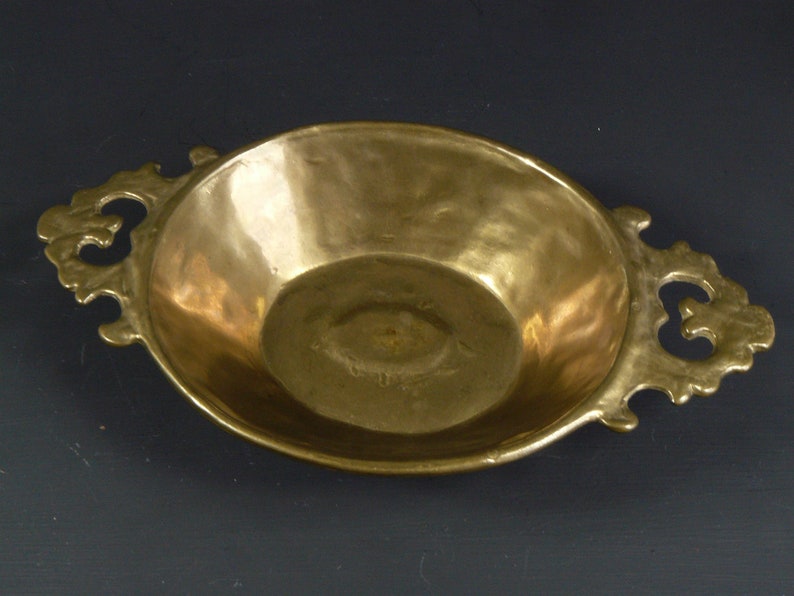 May include: A brass serving dish with two ornate handles. The dish is oval shaped and has a slightly raised rim.
