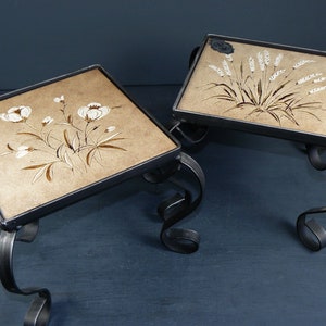 Pode incluir: Duas mesas pequenas, quadradas, de metal preto com tampos de cerâmica pintados à mão. Uma mesa apresenta um desenho floral com flores brancas e caules marrons. A outra mesa apresenta um desenho de trigo com espigas de trigo brancas e caules marrons.