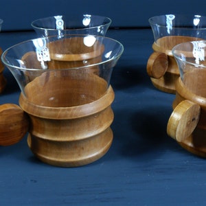 Op de afbeelding: Set van zes heldere glazen bekers met houten handvatten en bodems. De bekers hebben een uitlopende vorm en de handvatten zijn rond en hebben een gladde afwerking. De bodems zijn van hout en hebben een natuurlijke bruine kleur.