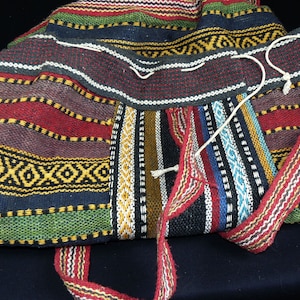 Op de afbeelding: Een kleurrijke geweven tas met een gestreept patroon in tinten rood, geel, blauw, groen en zwart. De tas heeft een lange band en is gemaakt van een dikke, duurzame stof.