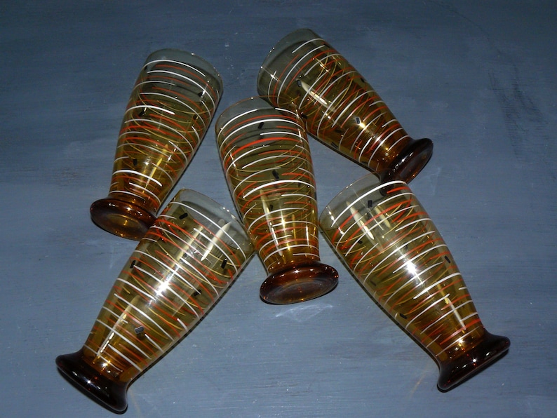 K&ouml;nnte beinhalten: Satz von f&uuml;nf Trinkgl&auml;sern aus Bernstein-Glas mit wei&szlig;en und orangefarbenen Streifenmustern. Die Gl&auml;ser haben einen abgerundeten Boden und einen ausgestellten oberen Rand.