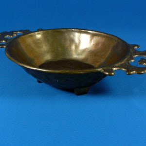May include: A brass serving bowl with ornate handles and three feet. The bowl is oval shaped and has a smooth, polished surface.