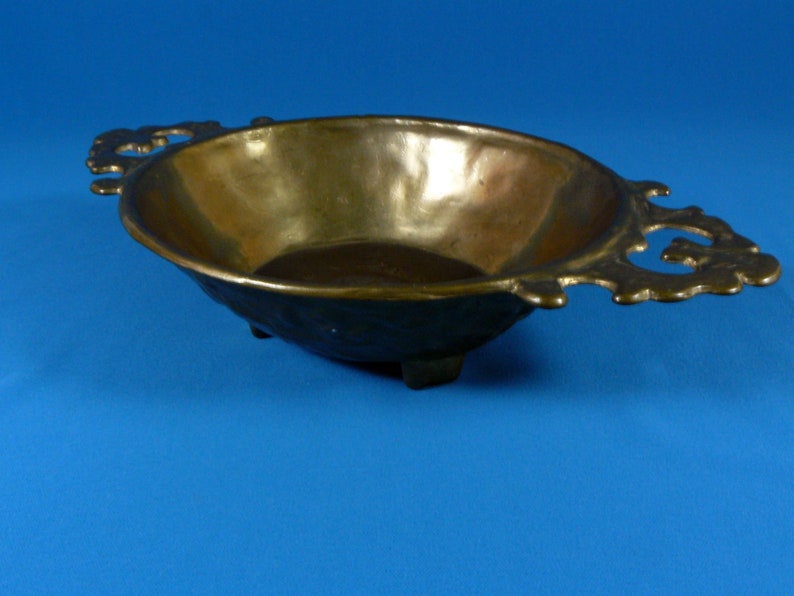 May include: A brass serving bowl with ornate handles and three feet. The bowl is oval shaped and has a smooth, polished surface.