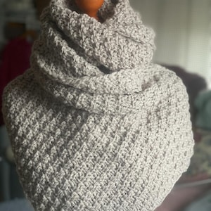 May include: A light gray, crocheted cowl scarf. The scarf is displayed on a mannequin head. The cowl is made of a textured stitch, and the fabric is folded over at the top. The scarf is a neutral color and appears soft and warm.
