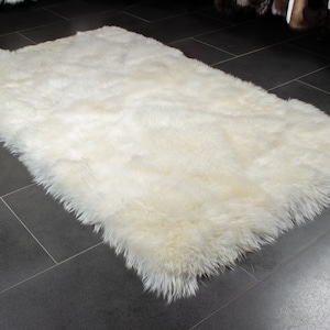Könnte beinhalten: Ein weißer Schaffellteppich mit weicher, flauschiger Textur. Der Teppich liegt auf einem dunkelgrauen Fliesenboden.